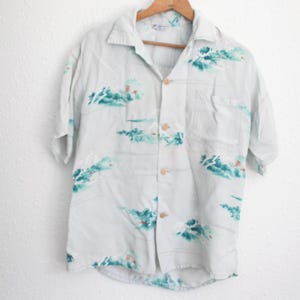 May include: A light blue button-down shirt with short sleeves, featuring a pattern of teal and white abstract shapes. The shirt is hanging on a wooden hanger against a white wall. The shirt has a single chest pocket and wooden buttons.