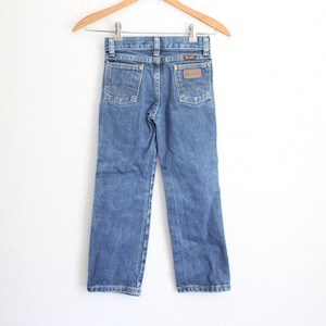 May include: Blue denim jeans hanging on a wooden hanger. The jeans have two back pockets with decorative stitching and a brown leather patch. The Wrangler brand label is visible on the back pocket. The jeans are a classic style.