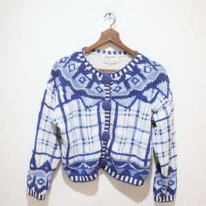 May include: A vintage blue and white cardigan sweater with a plaid and geometric pattern. The sweater has a round neckline, long sleeves, and large blue buttons. The sweater is hanging on a wooden hanger.