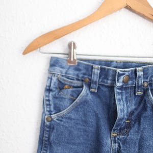 May include: Close-up of blue denim jeans hanging on a wooden hanger. The jeans have a classic five-pocket design with brass-coloured rivets and a button closure. The denim has a medium wash.