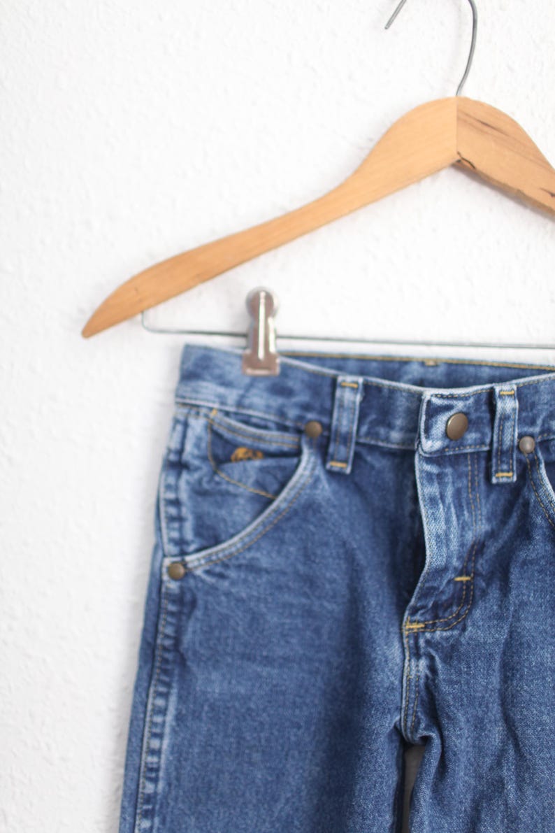May include: Close-up of blue denim jeans hanging on a wooden hanger. The jeans have a classic five-pocket design with brass-coloured rivets and a button closure. The denim has a medium wash.