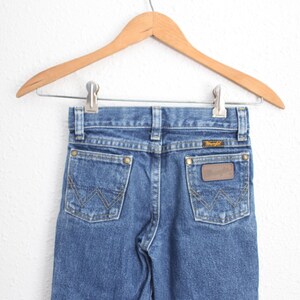May include: Blue denim jeans with the Wrangler logo on the back pocket, hanging on a wooden hanger. The jeans have classic five-pocket styling and a straight leg fit. The jeans are a medium wash.