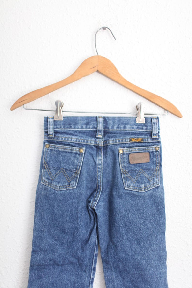May include: Blue denim jeans with the Wrangler logo on the back pocket, hanging on a wooden hanger. The jeans have classic five-pocket styling and a straight leg fit. The jeans are a medium wash.