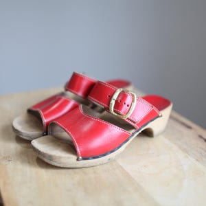 May include: A pair of red leather clogs with a buckle strap. The clogs have a wooden sole and are on a wooden surface.