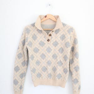 May include: A beige long-sleeve sweater featuring a light blue argyle diamond pattern. The garment has a collared neckline with a partial button-down placket containing two brown buttons and ribbed cuffs and hem.