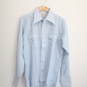 May include: Light blue button-down shirt with long sleeves, two chest pockets, and decorative embroidery near the shoulders. The shirt is hanging on a wooden hanger against a white wall. A vintage style shirt.