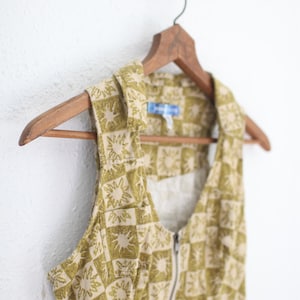 May include: A sleeveless vest with a sunburst pattern in olive green and beige, hanging on a wooden hanger. The vest has a zipper closure and a collared neckline. The fabric appears to be a lightweight cotton or linen.