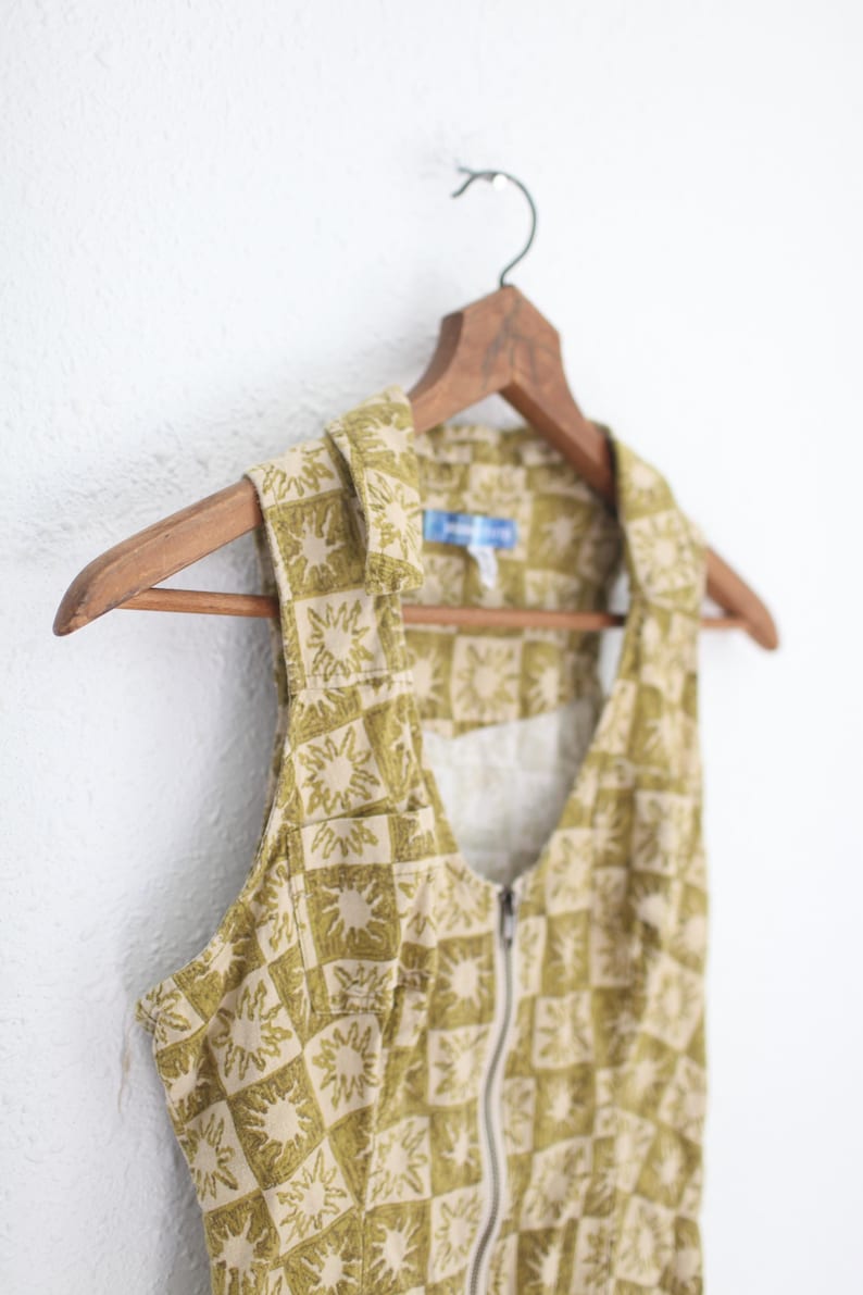 May include: A sleeveless vest with a sunburst pattern in olive green and beige, hanging on a wooden hanger. The vest has a zipper closure and a collared neckline. The fabric appears to be a lightweight cotton or linen.