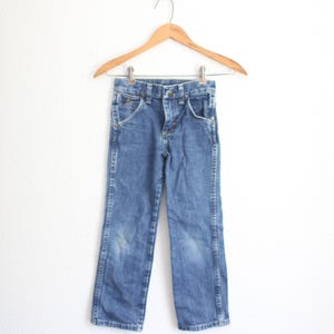 May include: Blue denim jeans hanging on a wooden hanger. The jeans have a classic five-pocket design, a button and zip closure, and a faded appearance. The jeans are likely for a child.