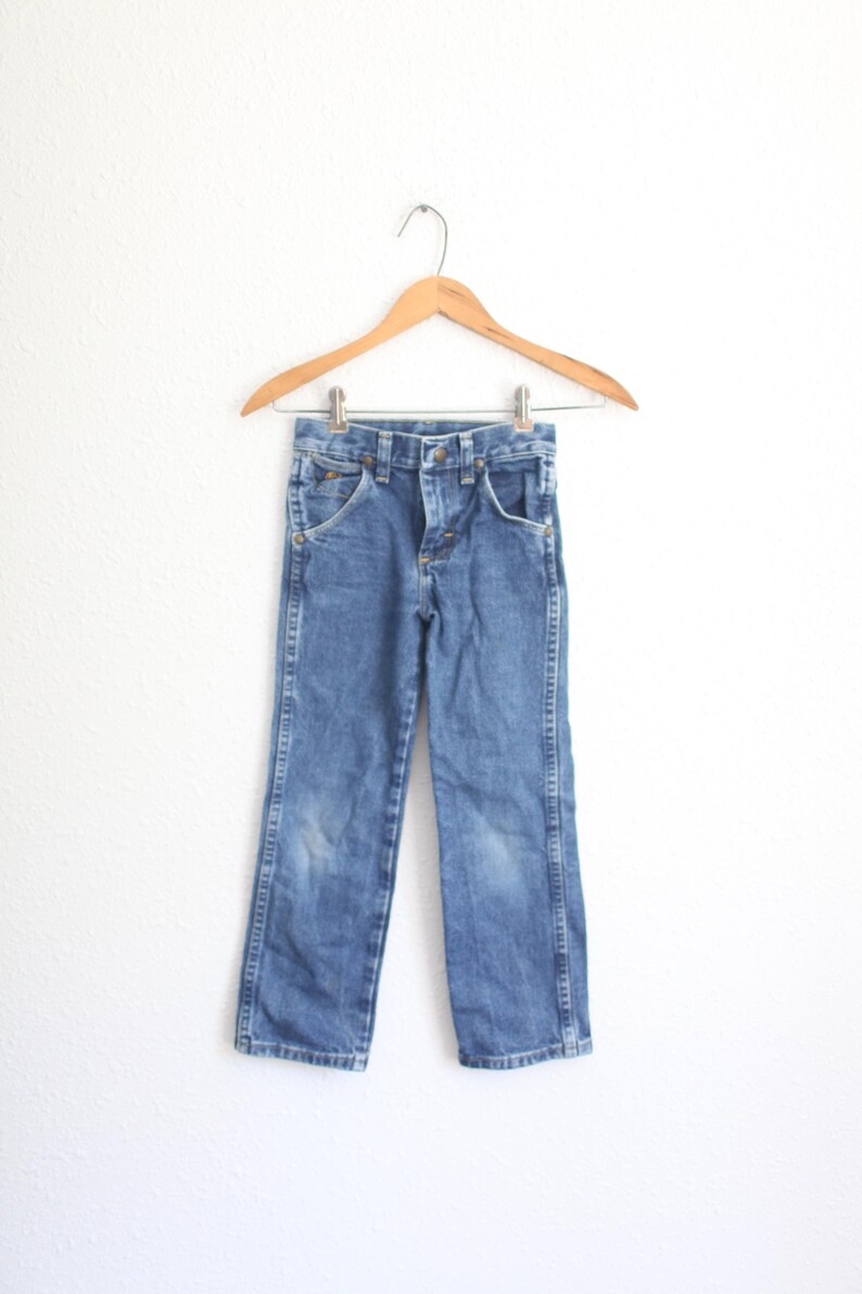 May include: Blue denim jeans hanging on a wooden hanger. The jeans have a classic five-pocket design, a button and zip closure, and a faded appearance. The jeans are likely for a child.