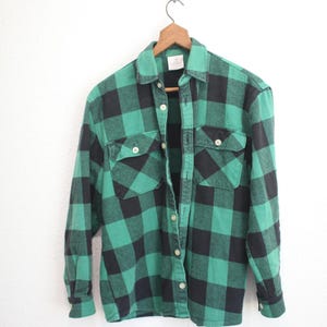 May include: Green and black plaid flannel shirt hanging on a wooden hanger. The long-sleeved shirt has two chest pockets and a button-down front. The plaid pattern is a classic design.
