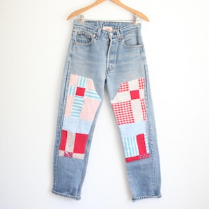 May include: Light blue denim jeans featuring large, rectangular patchwork panels on the front of each leg. The patches consist of various fabric squares in red, white, and light blue, displaying patterns such as gingham, stripes, and small floral prints.