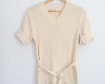 Pode incluir: Uma blusa de malha cor de creme de manga curta com decote em V, mangas com bainha e um cinto de tecido a condizer na cintura. A peça apresenta uma textura finamente canelada e é exibida num cabide de madeira contra uma parede branca.