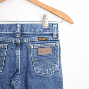 May include: Close-up of blue denim jeans with a brown leather patch and the Wrangler logo on the back pocket. The jeans are hanging on a wooden hanger. The jeans have a classic five-pocket design.