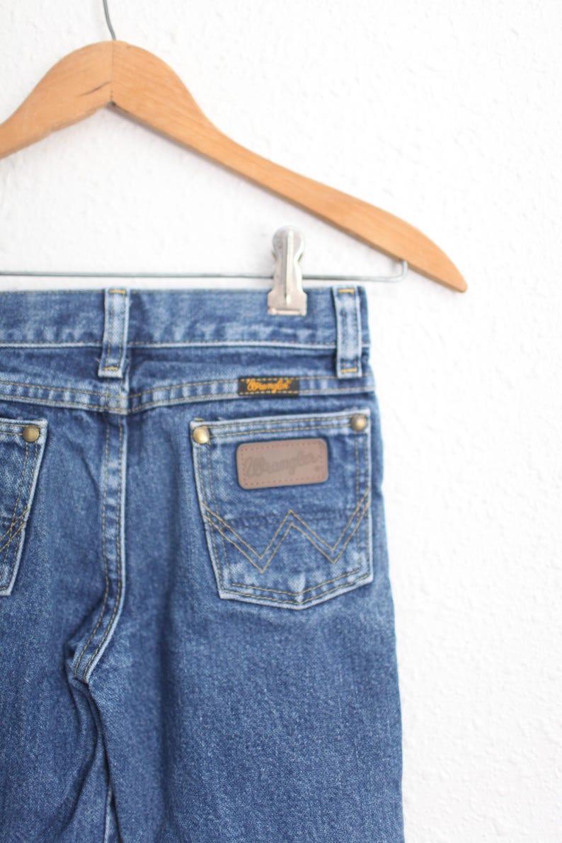 May include: Close-up of blue denim jeans with a brown leather patch and the Wrangler logo on the back pocket. The jeans are hanging on a wooden hanger. The jeans have a classic five-pocket design.