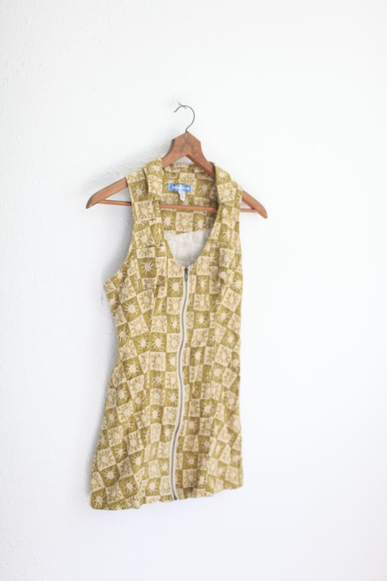 May include: A sleeveless, olive green dress with a repeating sunburst pattern in a lighter shade. The dress features a collared neckline, a full-length silver zipper, and hangs on a wooden hanger. The dress is a vintage style.