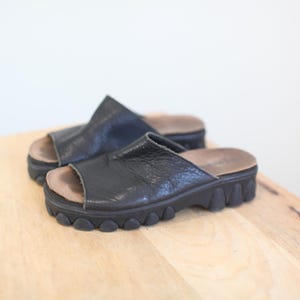 May include: A pair of black leather platform sandals with open toes. The sandals have a textured upper and a thick, ridged sole. The footbed is a light brown color. The shoes are placed on a light wooden surface.