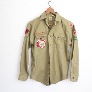 May include: Olive green button-down shirt with long sleeves, featuring multiple patches. Patches include the words "BOY SCOUTS OF AMERICA" and a circular patch with a mountain scene. The shirt is hanging on a wooden hanger.