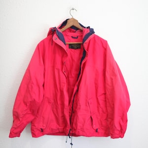 May include: A bright red hooded jacket with a navy blue trim and lining. The jacket has a drawstring at the waist and two front pockets. It is hanging on a wooden hanger against a white wall.