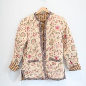 May include: A long-sleeve quilted jacket featuring a cream base with a multi-colored floral print in shades of pink, green, and brown. The jacket has a round neckline, a front button closure, two front pockets, and a contrasting patterned trim.