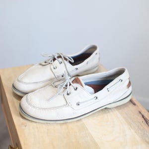 May include: A pair of white leather boat shoes with brown leather accents and white laces. The shoes are sitting on a light wood surface.