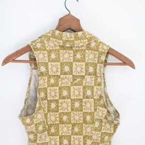 May include: A sleeveless top with a collared neckline, hanging on a wooden hanger. The top features a repeating pattern of olive green sunburst designs within a grid of beige squares. The fabric appears to be a lightweight material.