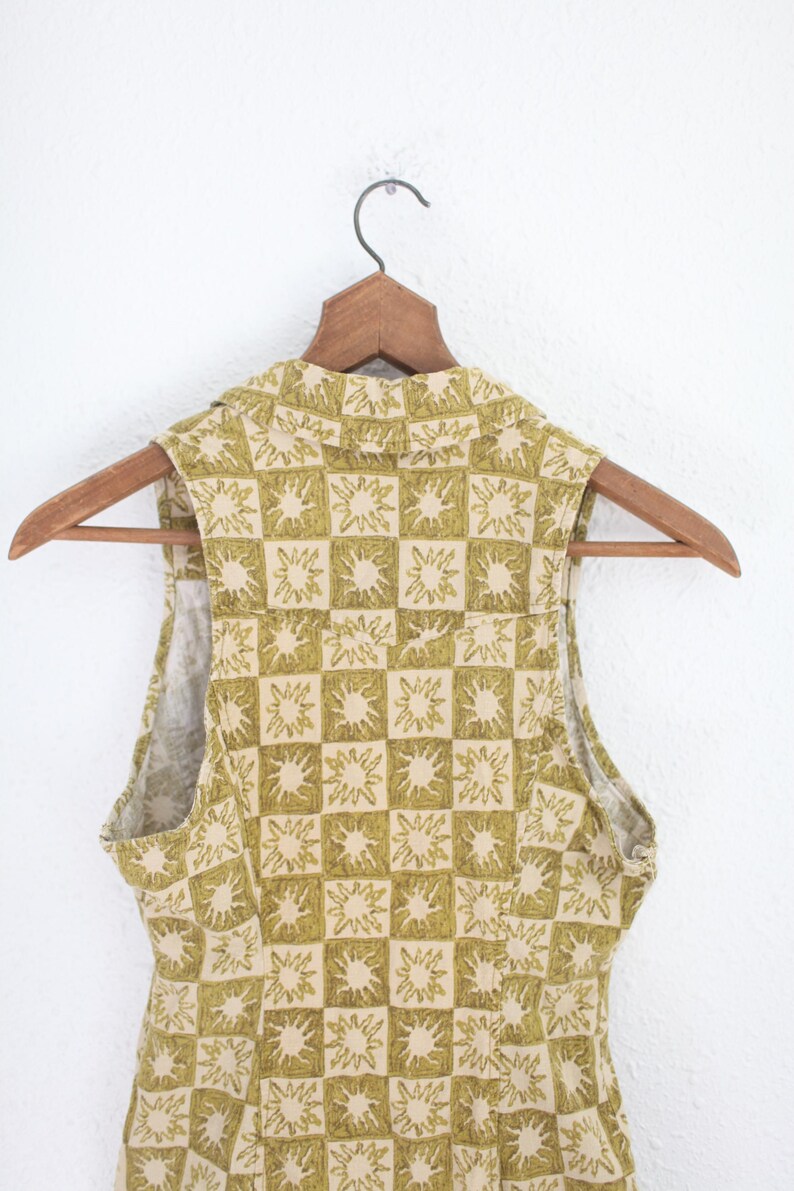 May include: A sleeveless top with a collared neckline, hanging on a wooden hanger. The top features a repeating pattern of olive green sunburst designs within a grid of beige squares. The fabric appears to be a lightweight material.