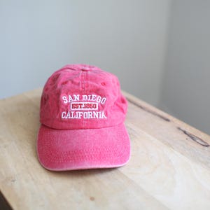 May include: A red baseball cap with the text "SAN DIEGO EST.1850 CALIFORNIA" embroidered in white. The cap is made of a soft material and sits on a wooden surface.