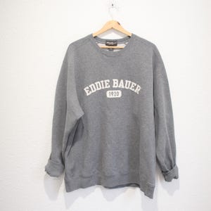 May include: A gray sweatshirt with the text "Eddie Bauer 1920" in white across the chest. The sweatshirt is hanging on a wooden hanger against a white wall. The sleeves are cuffed.