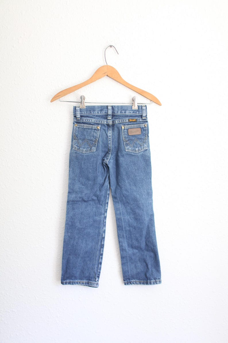 May include: Blue denim jeans hanging on a wooden hanger. The jeans have two back pockets with decorative stitching and a brown leather patch. The Wrangler brand label is visible on the back pocket. The jeans are a classic style.