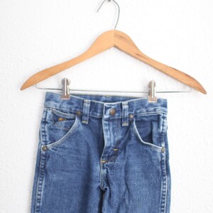 May include: Blue denim jeans hanging on a wooden hanger. The jeans feature classic five-pocket styling with a button and zip closure. The garment is made of a durable denim fabric, suitable for everyday wear.