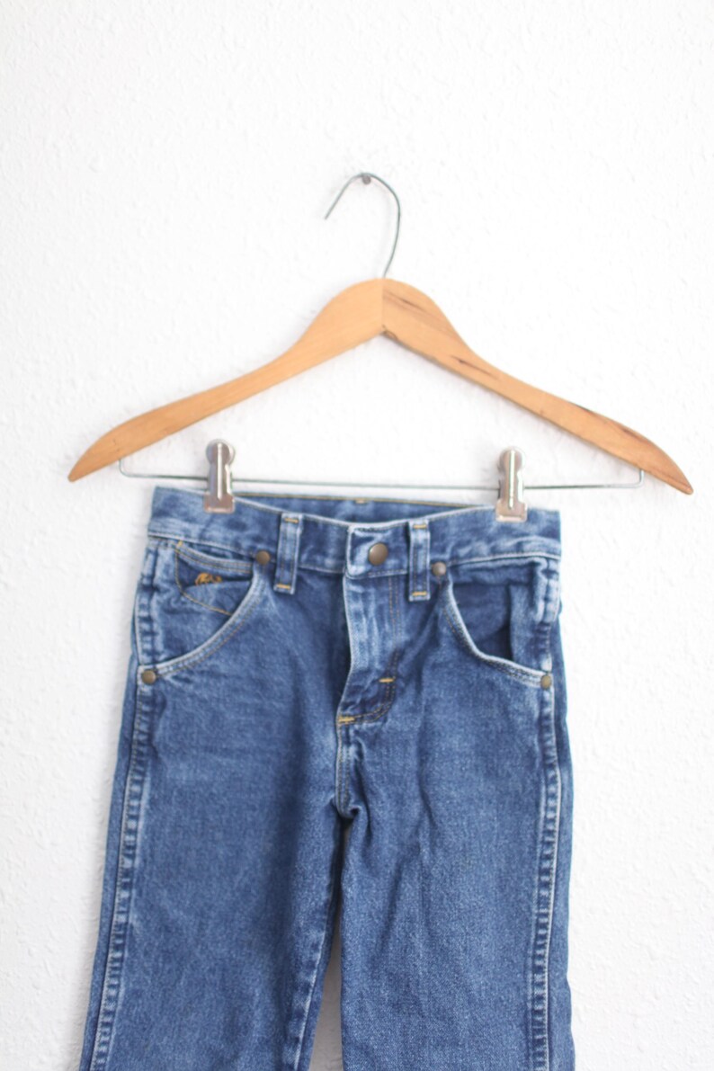 May include: Blue denim jeans hanging on a wooden hanger. The jeans feature classic five-pocket styling with a button and zip closure. The garment is made of a durable denim fabric, suitable for everyday wear.