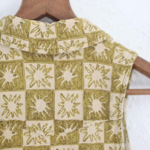 May include: Close-up of a sleeveless top with a unique pattern. The fabric features a repeating design of green sunbursts within a checkered pattern on a cream background. The top is hanging on a wooden hanger.
