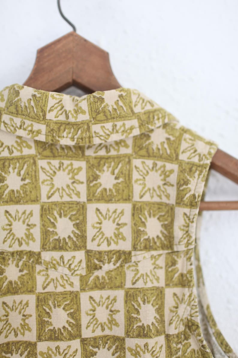 May include: Close-up of a sleeveless top with a unique pattern. The fabric features a repeating design of green sunbursts within a checkered pattern on a cream background. The top is hanging on a wooden hanger.