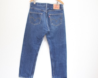 Puede incluir: Vaqueros azules de mezclilla colgados de una percha de madera. Los vaqueros tienen un diseño clásico de cinco bolsillos con una etiqueta roja en el bolsillo trasero. Los vaqueros son de lavado medio y parecen vintage.