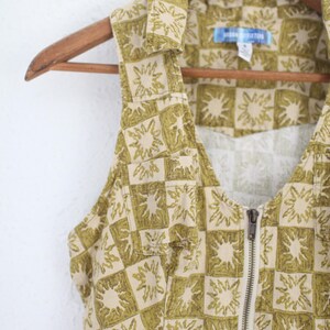 May include: A sleeveless, collared garment with a beige and olive green sunburst pattern. The garment has a full-length zipper and is hanging on a wooden hanger. The fabric has a checkered design.