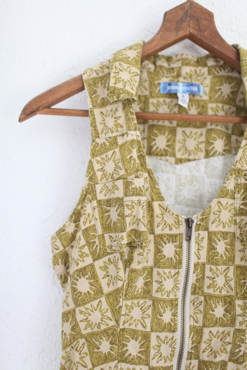 May include: A sleeveless, collared garment with a beige and olive green sunburst pattern. The garment has a full-length zipper and is hanging on a wooden hanger. The fabric has a checkered design.