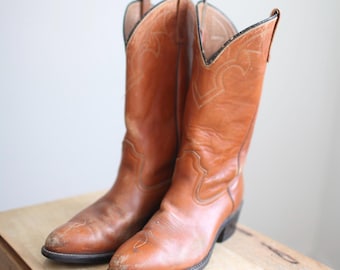 May include: A pair of brown leather cowboy boots with pointed toes and a slight heel. The boots have intricate stitching details on the shaft and a classic cowboy boot silhouette.
