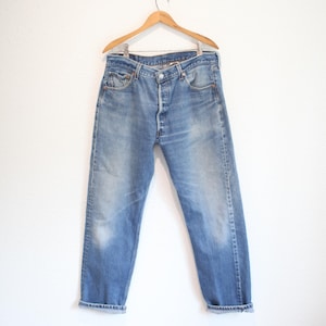 May include: A pair of medium-wash blue denim jeans featuring a button-fly closure, five-pocket styling, and a straight-leg cut. The fabric shows natural fading on the thighs and knees, with the cuffs rolled up at the bottom hem.