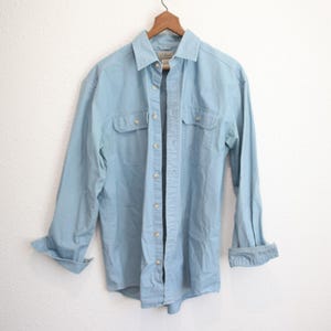May include: A light blue long-sleeve button-down shirt hanging on a wooden hanger. The shirt has two chest pockets and buttoned cuffs. The shirt is made of a soft, light-weight material.