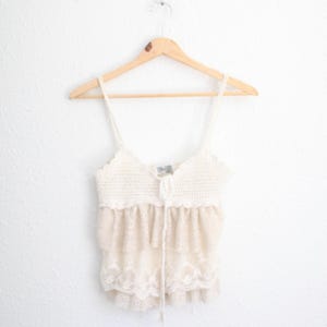 May include: Cream-colored crochet top with spaghetti straps and a tiered, lace-trimmed bottom. The top is hanging on a wooden hanger against a white wall. The top features a drawstring detail at the neckline.