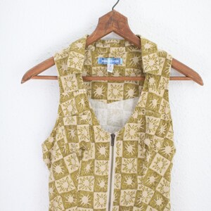 May include: Sleeveless dress with a collared neckline and a full-length zipper. The dress features a repeating pattern of olive green sunbursts on a tan background. The garment is hanging on a wooden hanger.