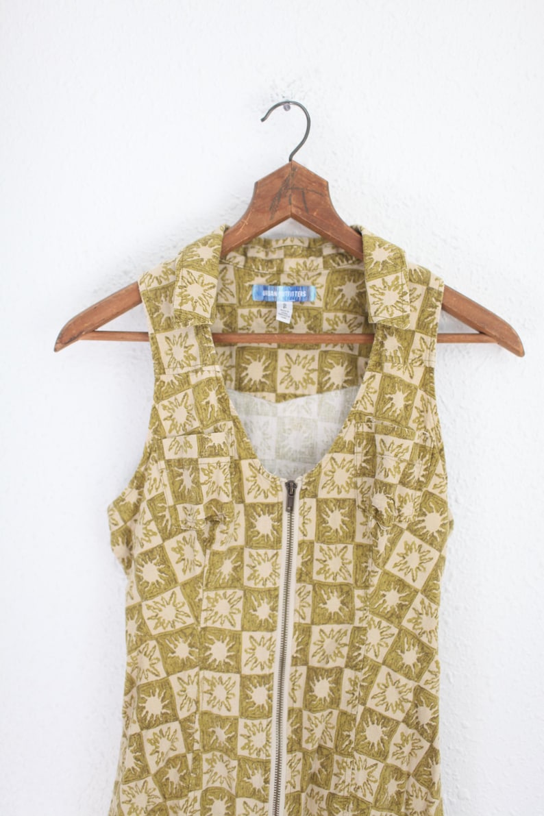 May include: Sleeveless dress with a collared neckline and a full-length zipper. The dress features a repeating pattern of olive green sunbursts on a tan background. The garment is hanging on a wooden hanger.