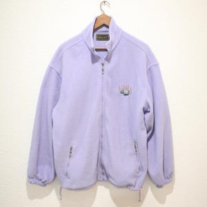 May include: A light purple fleece jacket with a full zipper closure and two side pockets. The jacket has a high collar and a drawstring at the hem. The word "Alaska" is embroidered on the chest.