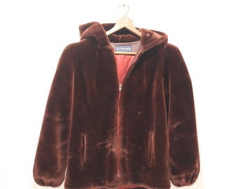May include: A brown faux fur hooded jacket with a full-length zipper and two front pockets. The jacket is hanging on a wooden hanger against a white background. The interior lining is red.
