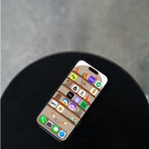 Puede incluir: Un teléfono inteligente negro con una pantalla blanca que muestra una cuadrícula de iconos de aplicaciones coloridos. El teléfono está sobre una superficie circular negra.