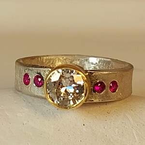May include: A silver and gold ring with a large, clear, round gemstone and four small, red gemstones.