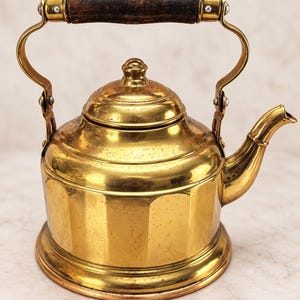 May include: A polished brass tea kettle featuring a rounded body with vertical fluted panels, a matching lid with a small knob, and a curved spout. The kettle has a hinged metal handle with a dark, weathered wooden grip secured by rivets.