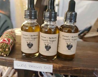 May include: Three glass bottles of Lucid Dream Oil with dropper tops. Each bottle has a label that reads "Stardust + Apothecary Starry Nights Lucid Dream Oil". The bottles are filled with a golden liquid.