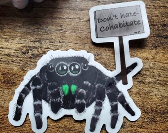 May include: A sticker of a jumping spider in black and white, with green eyes and a green patch. The spider holds a sign saying "Don't hate - Cohabitate". The sticker is on a wooden surface.